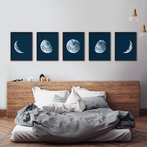 May include: Five framed prints of the moon in different phases, with a blue background. The prints are arranged in a row on a white wall above a wooden headboard.