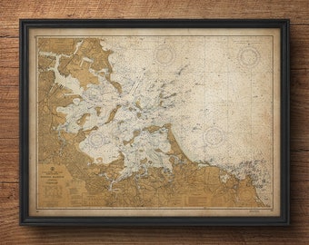 May include: A vintage nautical chart of Boston Harbour, printed on aged paper with a brown and cream colour scheme. The chart shows the coastline, islands, and waterways of the harbour, with detailed markings for depths, buoys, and other navigational features. The chart is framed in a black frame.