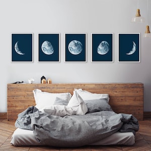 May include: Set of six blue and white moon phase prints on a dark blue background. The prints are arranged in a row on a white wall above a wooden headboard and a gray bed with white bedding.
