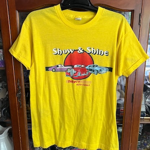 May include: Yellow short-sleeved t-shirt with a graphic of three cars and a red sun with the text "Show & Shine" and "Thrifty Auto Supply" printed on the front.