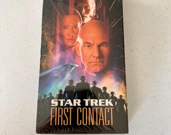 May include: A VHS tape of the film Star Trek: First Contact. The cover features a dark background with a futuristic spaceship and the main characters from the film, including Captain Jean-Luc Picard, Data, and the Borg Queen. The text "Resistance is Futile" is at the top of the cover, and the title "Star Trek First Contact" is in large, bold letters at the bottom.