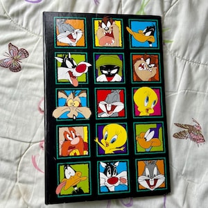 Op de afbeelding: Een zwarte en groene boekomslag met een raster van 16 kleurrijke vierkanten. Elk vierkant toont een ander cartoonpersonage uit de Looney Tunes-franchise, waaronder Bugs Bunny, Daffy Duck, Tweety Bird en Sylvester de Kat.