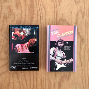 Op de afbeelding: Twee cassettebandjes met roze en blauwe hoezen. De cassette aan de linkerkant is getiteld "Timepieces: The Best of Eric Clapton". De cassette aan de rechterkant is getiteld "Eric Clapton: Too Much Monkey Business".