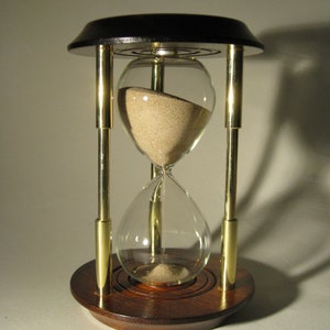 May include: A wooden hourglass with brass accents and a glass bulb filled with sand. The hourglass is on a white surface.