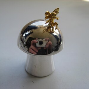 Könnte beinhalten: Silberfarbene, pilzförmige Dekoration mit einer goldfarbenen Feenfigur. Der Pilz hat eine glänzende, reflektierende Oberfläche. Der Sockel ist weiß. Die Fee ist detailliert mit Flügeln und einer skurrilen Pose.