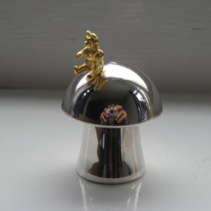 Könnte beinhalten: Ein silbernes, pilzförmiges Deko-Objekt mit einer goldfarbenen Figur obenauf. Der Pilz hat eine glänzende, reflektierende Oberfläche. Die Figur scheint eine kleine Person oder Kreatur zu sein.