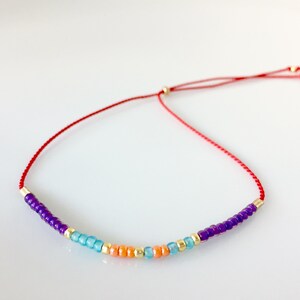 May include: A red string bracelet with purple, blue, and orange beads. The bracelet is tied with a knot and has a gold bead at each end.