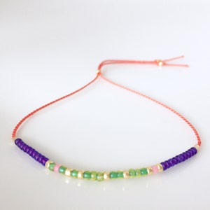 May include: A red string bracelet with purple, green, and pink beads. The beads are small and round, and the string is thin.
