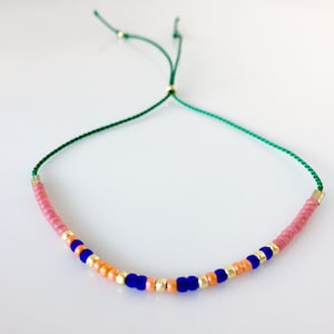 May include: A green string bracelet with pink, orange, and blue beads. The bracelet is adjustable with a sliding knot.