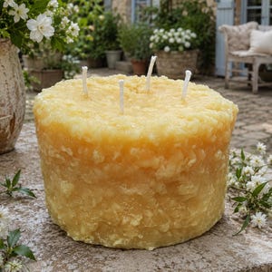 May include: A large, cylindrical, golden-yellow beeswax candle with a textured, granular surface. The candle features four white cotton wicks protruding from the top. The wax has a semi-translucent appearance with visible crystalline patterns.