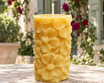 May include: A cylindrical beeswax candle with a golden yellow hue, featuring a detailed relief pattern of multiple blooming flowers covering its entire exterior surface and a single white cotton wick centered at the top.