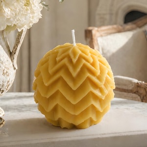May include: A yellow, spherical candle with a geometric, zig-zag pattern. The candle sits on a white rectangular tray on a marble table. A white floral arrangement in a distressed white vase is in the background.