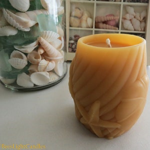 May include: A large, hand-crafted, yellow beeswax candle with a starfish design. The candle is sitting on a white surface next to a glass jar filled with seashells and sea glass.