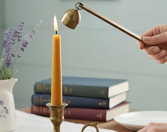 May include: A lit, slender, yellow candle in a brass candlestick is being extinguished with a brass candle snuffer. The candle snuffer has a bell-shaped cover attached to a long handle. A white pitcher with blue floral designs and a framed picture are in the background.