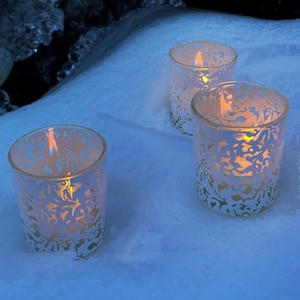 Peut inclure: Trois bougeoirs en verre blanc avec un motif floral délicat sont allumés et placés sur une surface enneigée. Les bougies brillent de mille feux, créant une ambiance chaleureuse et accueillante. Le texte "Bees Light Candles®" est visible en arrière-plan.