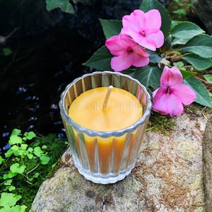 May include: A small, round, yellow beeswax candle in a clear glass jar with a ribbed design. The candle is sitting on a rock with green moss and pink flowers in the background.
