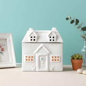 May include: A white ceramic house-shaped wax warmer with a removable roof, windows, and a door. The warmer has a chimney and is decorated with small flower pots. The background is a light blue wall.