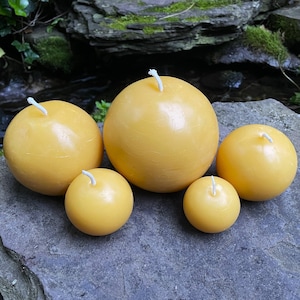 May include: Five round, yellow beeswax candles of varying sizes. The candles are arranged on a grey rock surface.