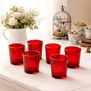 May include: Six red drinking glasses are arranged on a white lace tablecloth. The glasses are cylindrical with a thick base. A white pitcher with flowers, a decorative birdcage, and small potted plants are in the background.