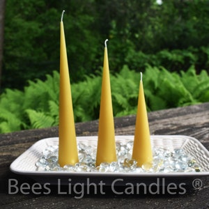May include: Three tapered beeswax candles of varying sizes, arranged on a white tray with clear glass stones. The candles are a light yellow color and have white wicks. The tray is on a wooden surface. The text "Bees Light Candles" is visible below the tray.