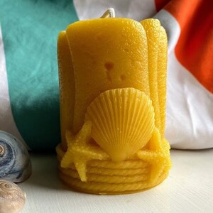 Op de afbeelding: Een gele bijenwaskaars met een schelp- en touwontwerp. De kaars staat op een wit oppervlak met twee schelpen.