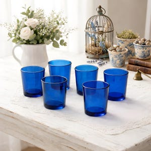 May include: Six cobalt blue drinking glasses are arranged on a white lace tablecloth. The glasses are cylindrical with a thick base and a simple, elegant design. The scene includes a white pitcher with flowers and decorative items.