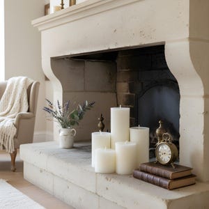 May include: A collection of ivory pillar candles of varying heights arranged on a stone fireplace mantel. A small brass clock and two leather-bound books sit nearby. A vase of lavender and greenery adds a touch of color.