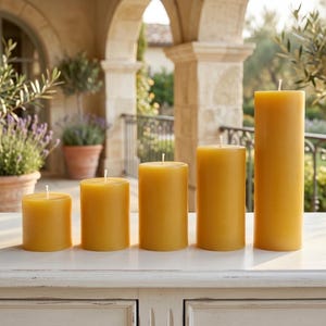 May include: Five yellow pillar candles of varying heights are arranged in a row. The candles are cylindrical with a smooth surface and a wick at the top. The background features an outdoor setting with architectural details and greenery.