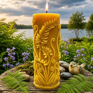 May include: A lit, yellow beeswax candle with a detailed botanical design. The candle features carved leaves and floral patterns. The candle is placed on a wooden surface with greenery and a small bird figurine in the background.