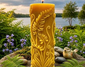 May include: A lit, yellow beeswax candle with a detailed botanical design. The candle features carved leaves and floral patterns. The candle is placed on a wooden surface with greenery and a small bird figurine in the background.