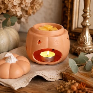 May include: A terracotta pumpkin-shaped wax warmer featuring star-shaped cutouts and a top basin holding yellow wax melts. A lit tea light candle sits in the lower opening. Beside the warmer rests a matching pumpkin-shaped lid with a white stem.