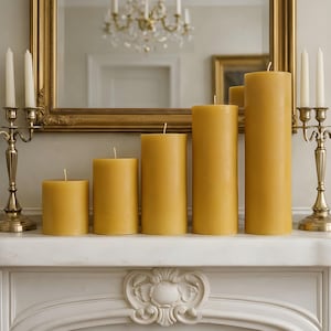 May include: Five cylindrical yellow beeswax pillar candles of varying heights arranged in a row. The candles have a smooth, solid texture and a single centered cotton wick each. They are positioned on a flat surface between two brass candle holders.