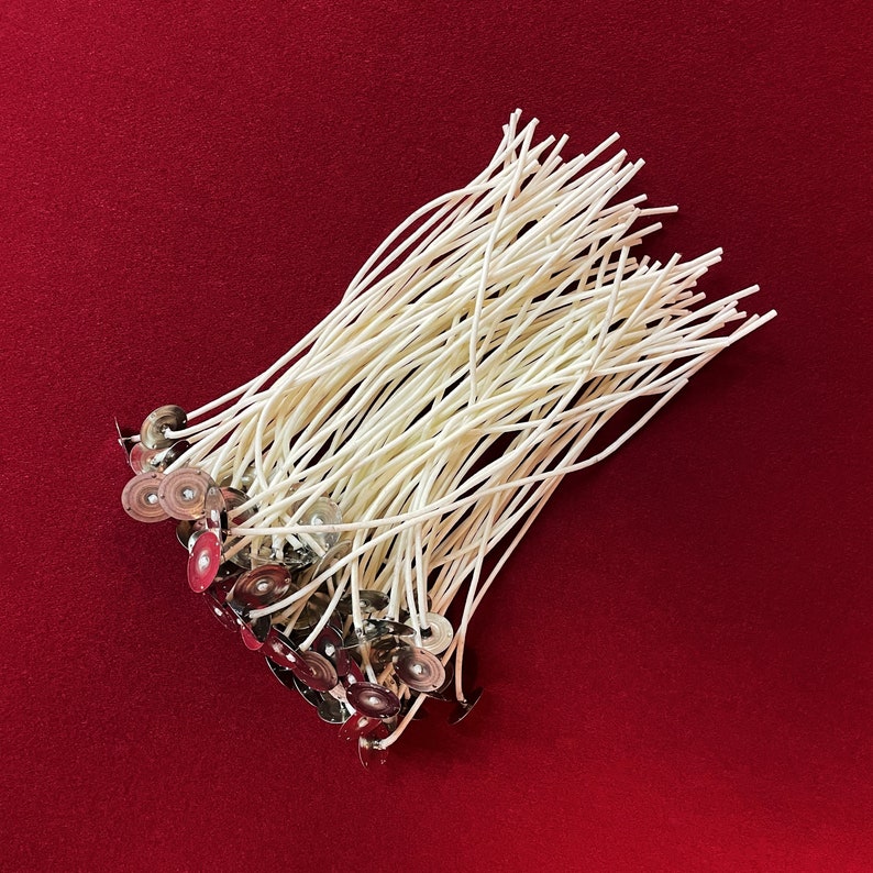 May include: A bundle of white cotton candle wicks with metal bases. The wicks are arranged in a pile on a red surface.