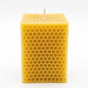 May include: A square, yellow beeswax candle with a honeycomb pattern. The candle has a white wick centered on the top. The candle is a solid block of wax, with a textured surface. The candle is a decorative item.