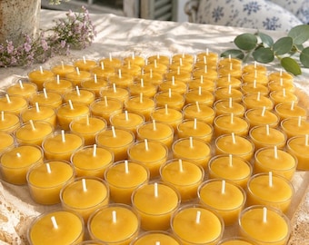 May include: A collection of yellow tea light candles arranged in clear plastic cups. Each candle has a white wick centered in the wax. The candles are on a white lace-trimmed surface, with flowers in the background.