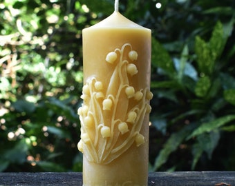 May include: A pillar candle in a warm yellow colour, decorated with a detailed relief of lily of the valley flowers. The candle has a central wick and is set against a blurred green backdrop. The base of the candle has the text "LightCandles".