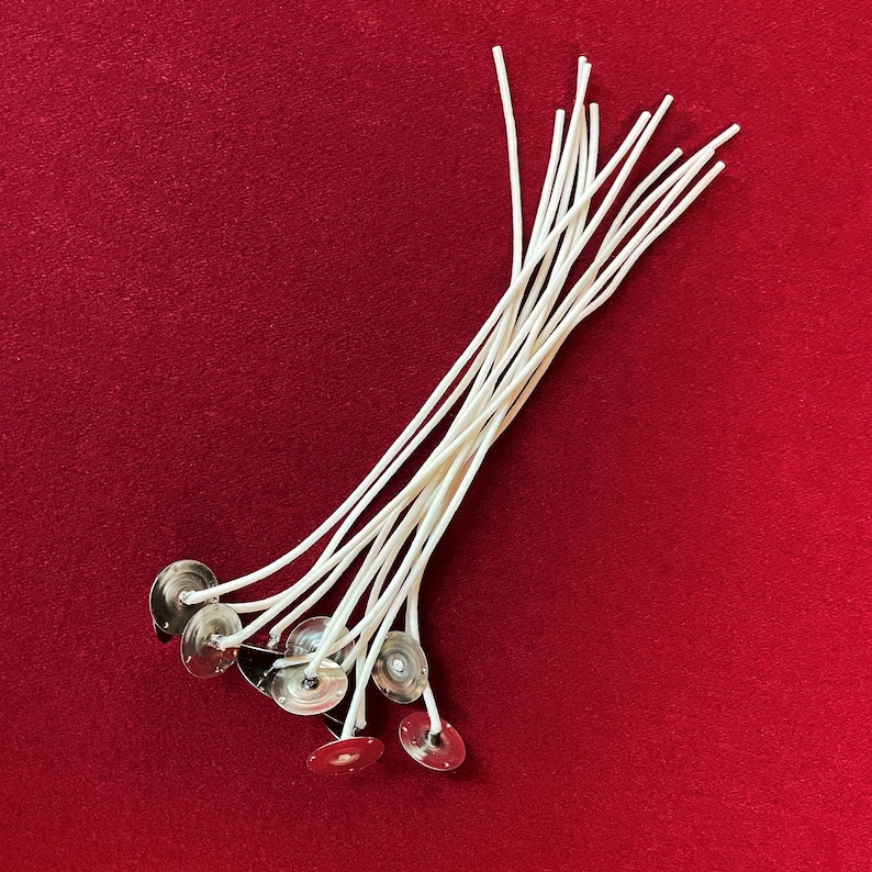 May include: A bundle of white cotton candle wicks with metal bases. The wicks are arranged in a fan shape on a red surface.