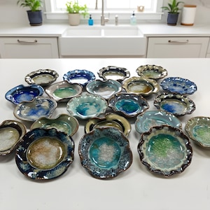 May include: A collection of handmade ceramic dishes with unique, organic shapes and glazed finishes. The dishes feature a variety of colors, including blues, greens, and browns, with textured rims. The arrangement showcases the variety of designs.