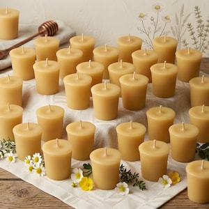 May include: A collection of unscented, light yellow votive candles arranged on a white cloth, with a honey dipper in the background. The candles are surrounded by small white and yellow flowers and greenery, creating a warm, inviting ambiance.
