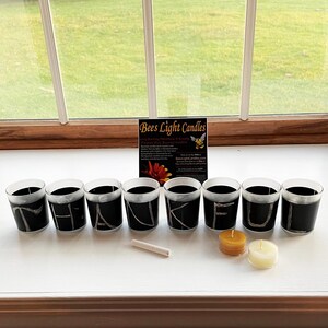 May include: A collection of black votive candle holders with white lettering spelling out a word. Two small candles and a stick of incense are also present. A sign for "Bees Light Candles" is in the background.