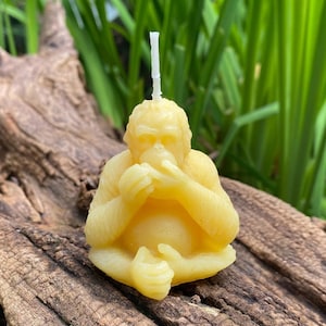May include: A hand-crafted beeswax candle shaped like an orangutan sitting with its hands clasped in front of it. The candle is a light yellow color and has a white wick.