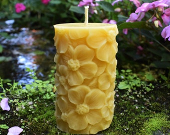 May include: A yellow beeswax candle with a floral design. The candle is shaped like a cylinder and has a white wick. The candle is sitting on a bed of moss.