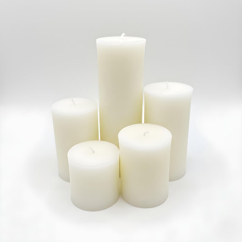 Puede incluir: Cinco velas de pilar de color marfil de diferentes alturas dispuestas sobre una superficie blanca. Las velas sin perfume tienen una forma cil&iacute;ndrica lisa. Cada vela tiene una mecha en el centro.