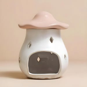 Könnte beinhalten: Ein weißer, pilzförmiger Ölverdampfer aus Keramik mit hellrosa Kappe. Der Verdampfer hat eine große Öffnung und rautenförmige Aussparungen. Der Innenraum ist dunkelgrau, vor einem hellpfirsichfarbenen Hintergrund.