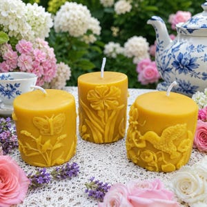 May include: Three cylindrical yellow beeswax candles featuring embossed nature designs. The left candle shows a butterfly and lavender, the center candle displays a large flower, and the right candle depicts a hummingbird among flowers.