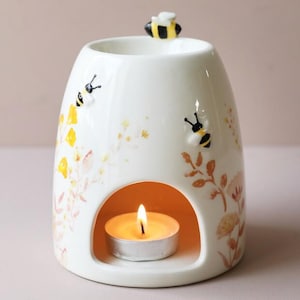 Könnte beinhalten: Ein weißer Keramik-Diffusor für ätherische Öle mit einem Bienen- und Blumendesign. Der Diffusor hat eine runde Öffnung für eine brennende Teelichtkerze. Der Diffusor ist mit gelben und orangen Blumen und zwei kleinen Bienenfiguren verziert.
