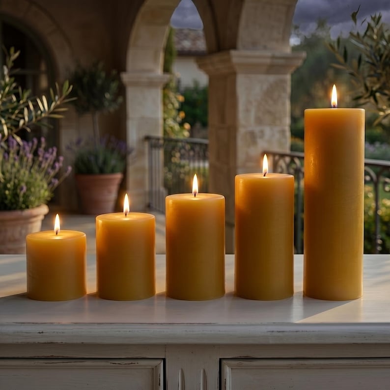 May include: Five lit, golden-yellow pillar candles of varying heights are arranged in a row. The candles are cylindrical and have a smooth surface. The flames are bright and create a warm, inviting ambiance. The candles are on a white surface.