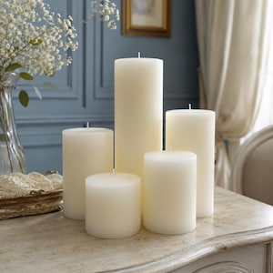 Puede incluir: Cinco velas de pilar de color marfil de diferentes alturas se colocan sobre una superficie clara. Las velas son cil&iacute;ndricas, lisas y tienen una mecha en la parte superior. El fondo incluye un jarr&oacute;n con flores blancas y una pared azul.