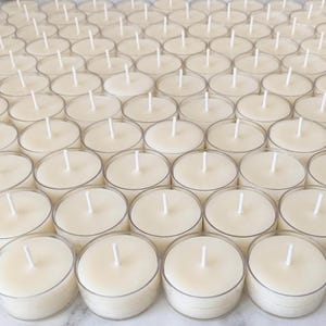 May include: An overhead shot of numerous tea light candles arranged in a grid pattern. Each candle is cream-colored with a white wick centered in a clear plastic cup. The candles are evenly spaced, creating a symmetrical and organized display.