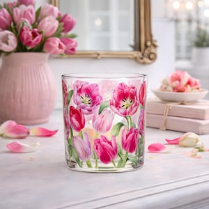 May include: A clear glass candle holder decorated with pink tulip flowers. The candle holder is cylindrical and sits on a white surface. A pink vase with tulips and a mirror are in the background.